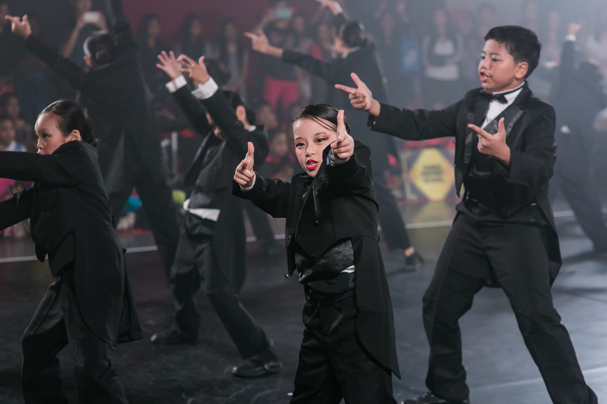Children in black tuxedos perform a coordinated dance on stage before a crowd.